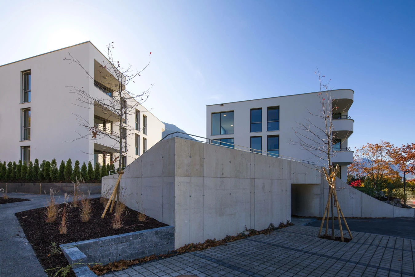 Wohnanlage Wolf-Huber-Straße in Feldkirch mit weißen Gebäuden, Tiefgarageneinfahrt, begrünten Beeten und gepflastertem Gehweg an einem sonnigen Tag.