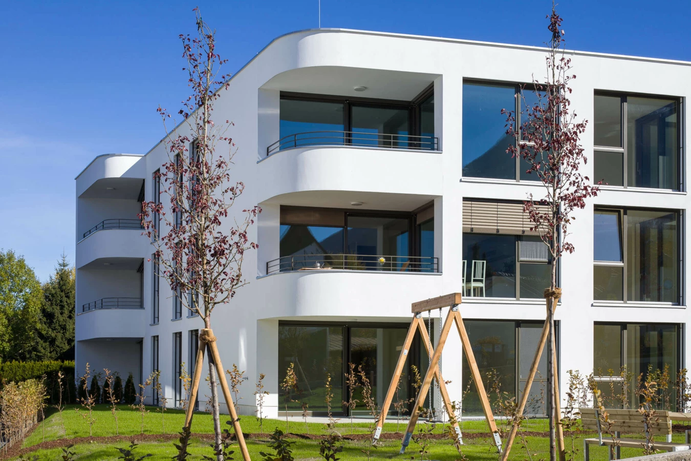 Modernes, weisses Wohngebäude mit geschwungenen Balkonen und großen Fenstern, umgeben von Grünflächen und jungen Bäumen (Wolf-Huber-Straße, Wohnanlage, Feldkirch).