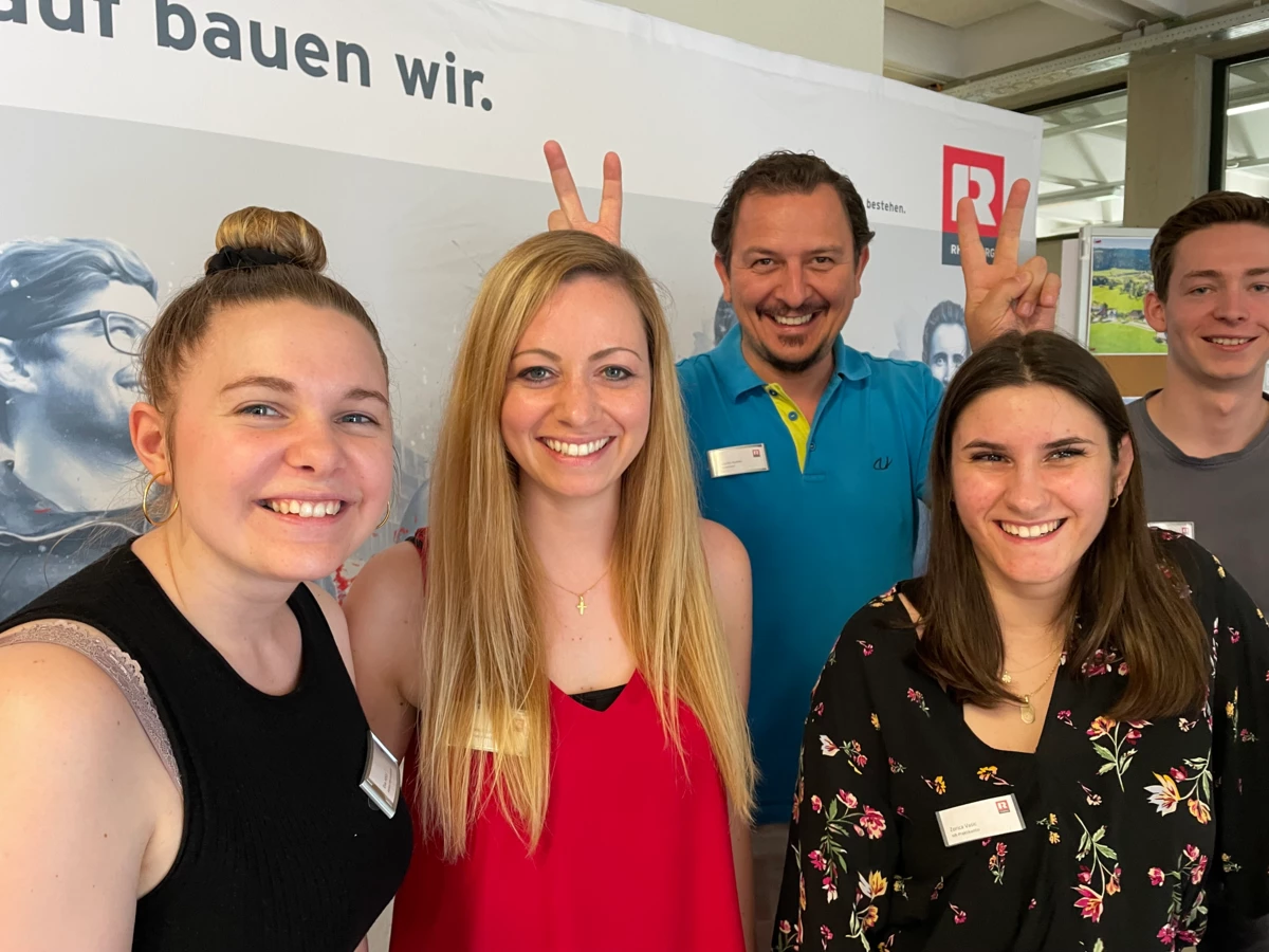 Eine Gruppe fröhlicher junger Erwachsener posiert lachend vor einem Rhomberg Bau Banner. Ein Mann macht Hasenohren hinter einer Frau (Bauforum).