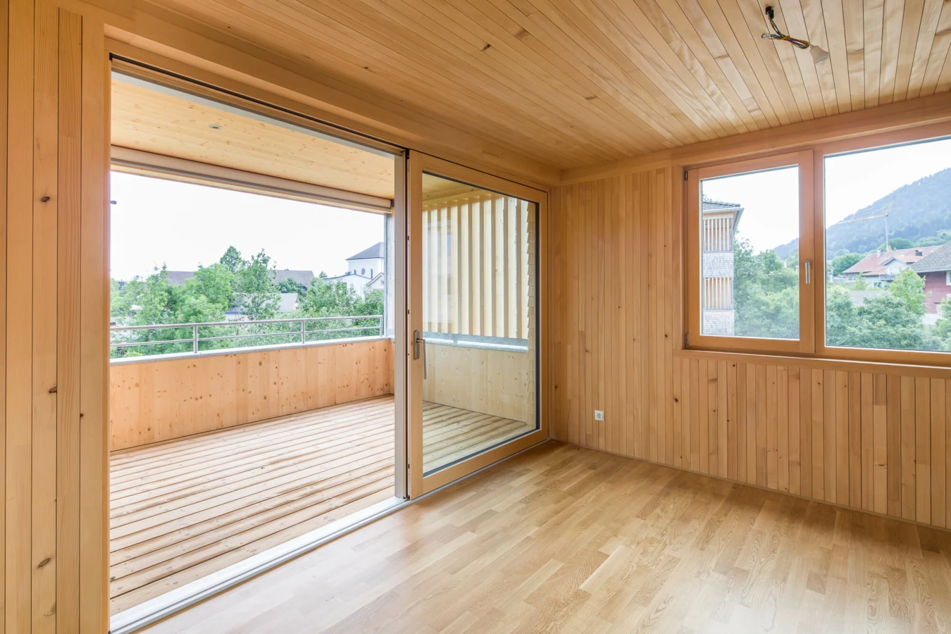 Ein Zimmer mit hellen Holzpaneelen an Wänden und Decke, Holzboden, Schiebetür zum Balkon und Fenster mit Blick ins Grüne (Wohnanlage, Alberschwende).