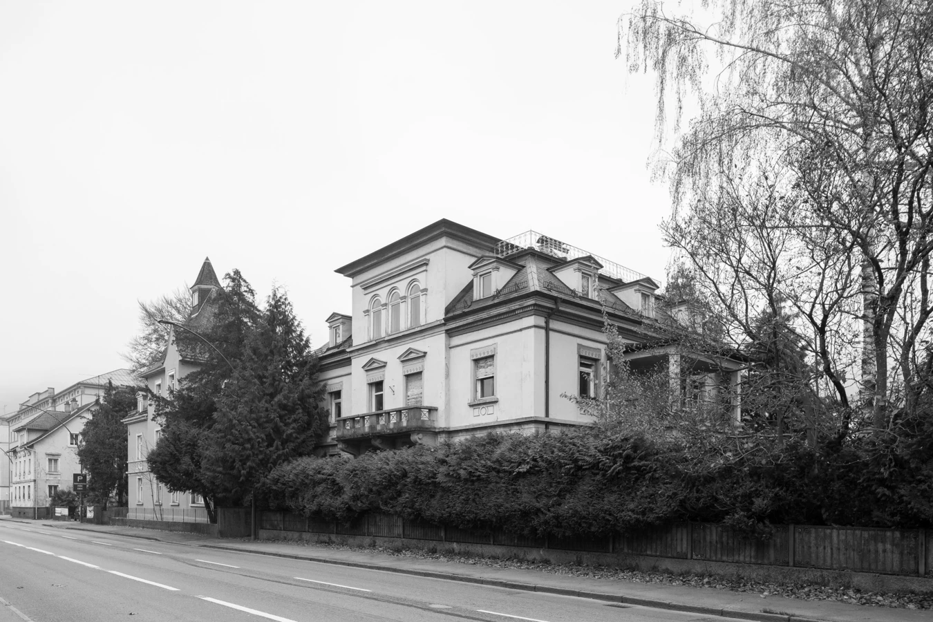 Schwarz-Weiß-Ansicht der Villa Reichsstraße, einer historischen Residenz mit großem Balkon hinter einer hohen Hecke, an einer Straße in Bregenz.