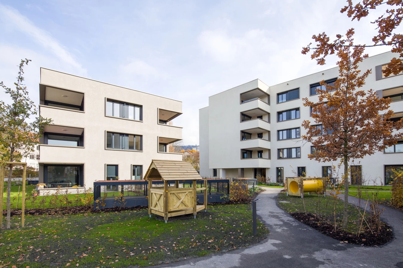 Zwei moderne weiße Wohngebäude mit Balkonen, umgeben von einem begrünten Hof mit Spielhaus und Tunnel (Bäumleareal)