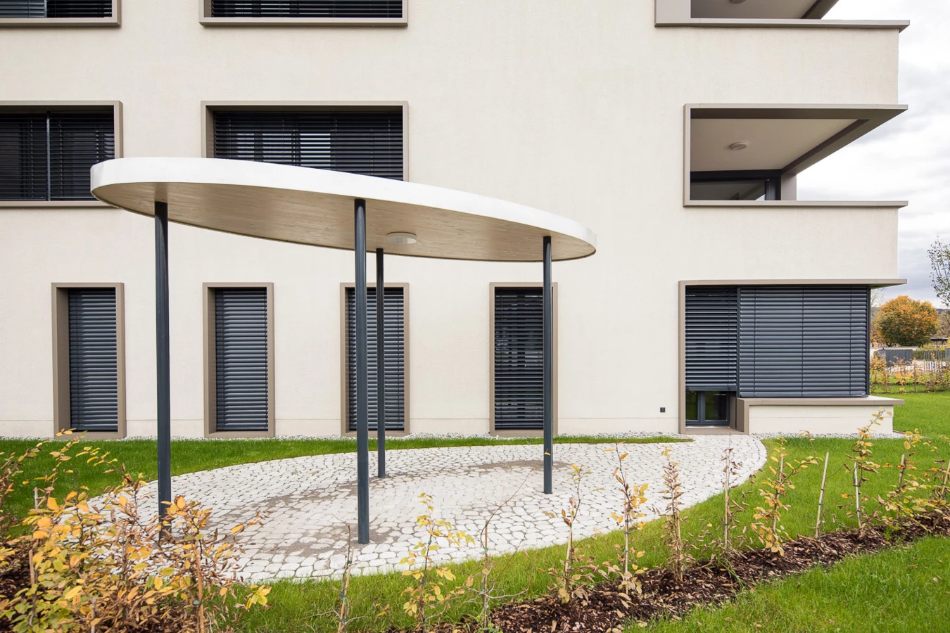 Eingangsbereich eines modernen Gebäudes mit ovalem Vordach, gepflastertem Weg und Grünflächen mit jungen Sträuchern (Baeumlequartier).