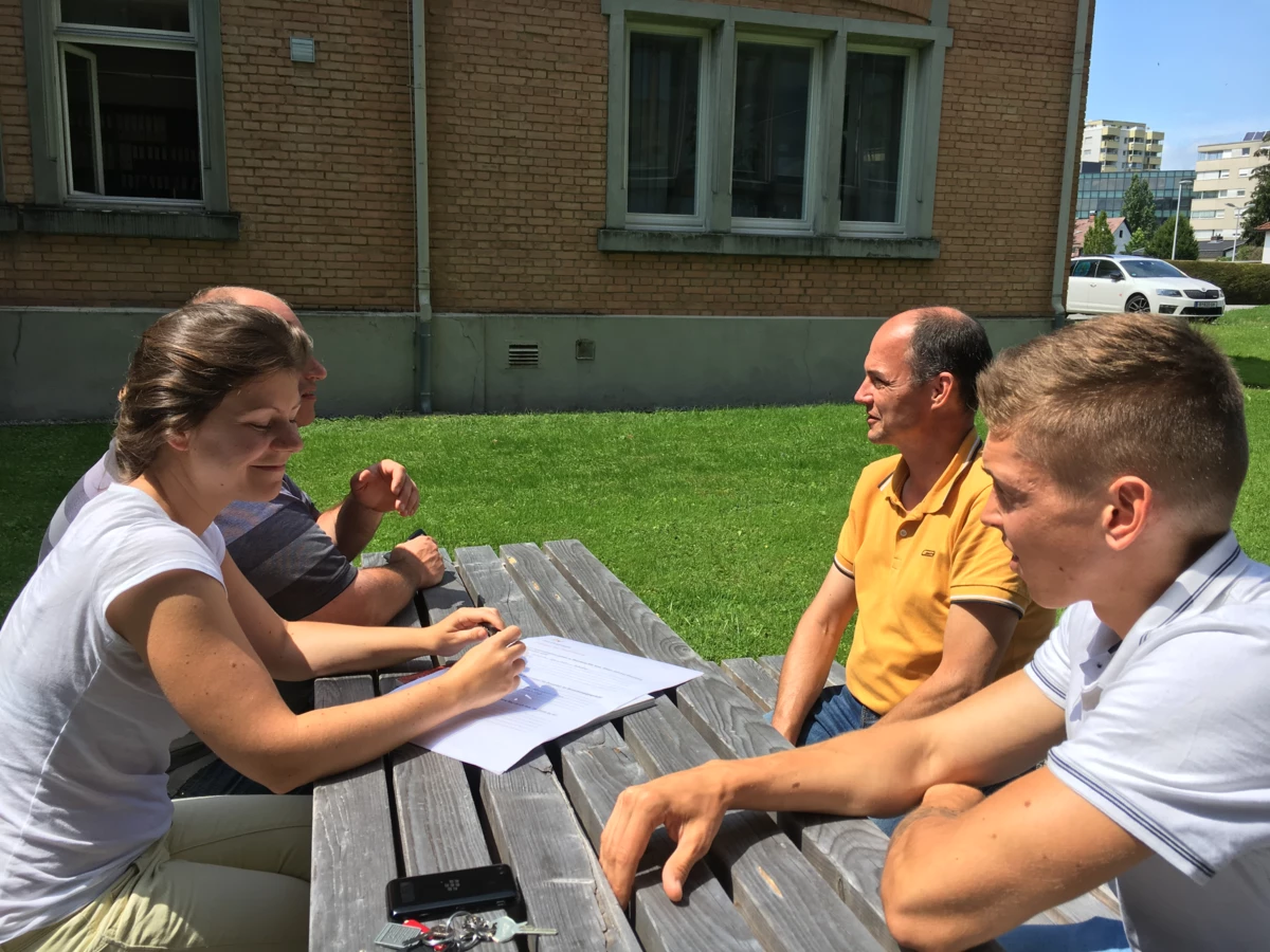 Vier Personen sitzen bei sonnigem Wetter an einem Holztisch im Freien, unterhalten sich und lesen Dokumente (Workshop).