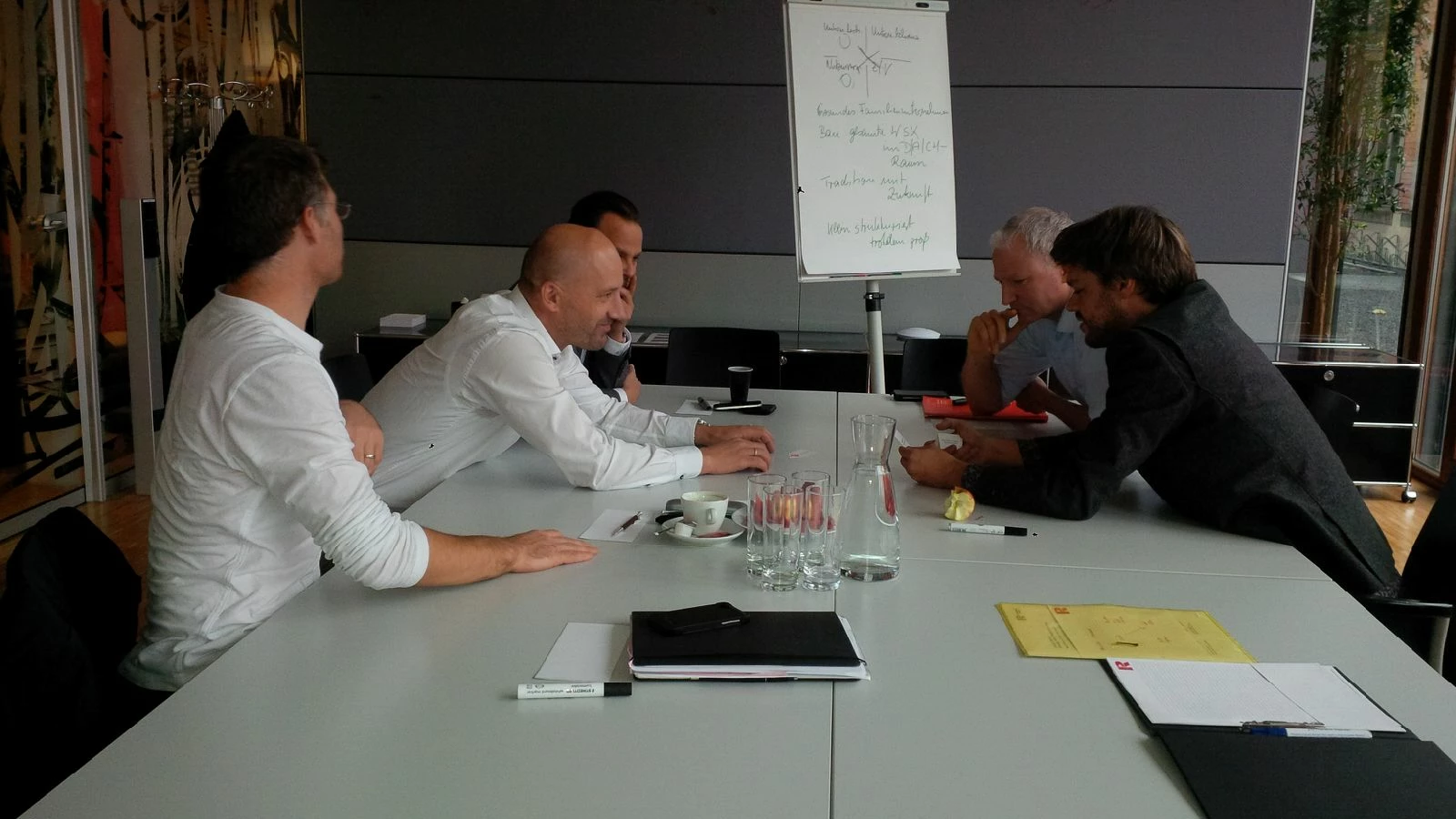 Fünf Männer sitzen an einem Konferenztisch mit Unterlagen und einem Flipchart im Hintergrund, bei einem Workshop (Führungskräfte).