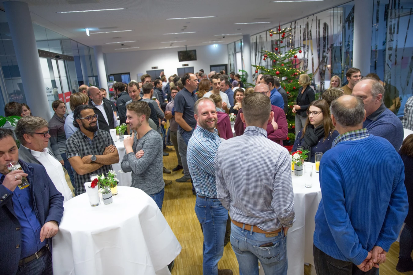 Eine große Gruppe von Menschen unterhält sich informell in einem hellen, modernen Raum mit einem Weihnachtsbaum, Stehtischen und Getränken (Hubert, Geburtstag).