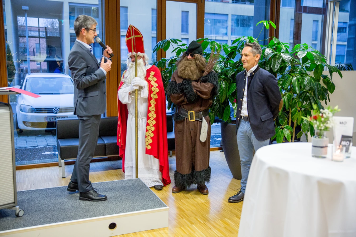 Ein Mann im Anzug spricht ins Mikrofon neben einem als Nikolaus verkleideten Mann, einem Mann in dunkler Kleidung mit Bart und einem Mann im Trachtenjanker (Hubert, Geburtstag).
