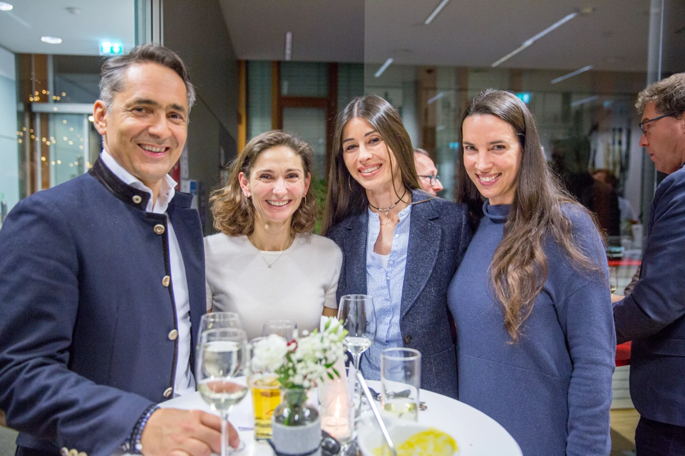 Vier lachende Personen, drei Frauen und ein Mann in Trachtenjacke, stehen an einem Stehtisch mit Getränken bei einer Veranstaltung (Hubert, Geburtstag).