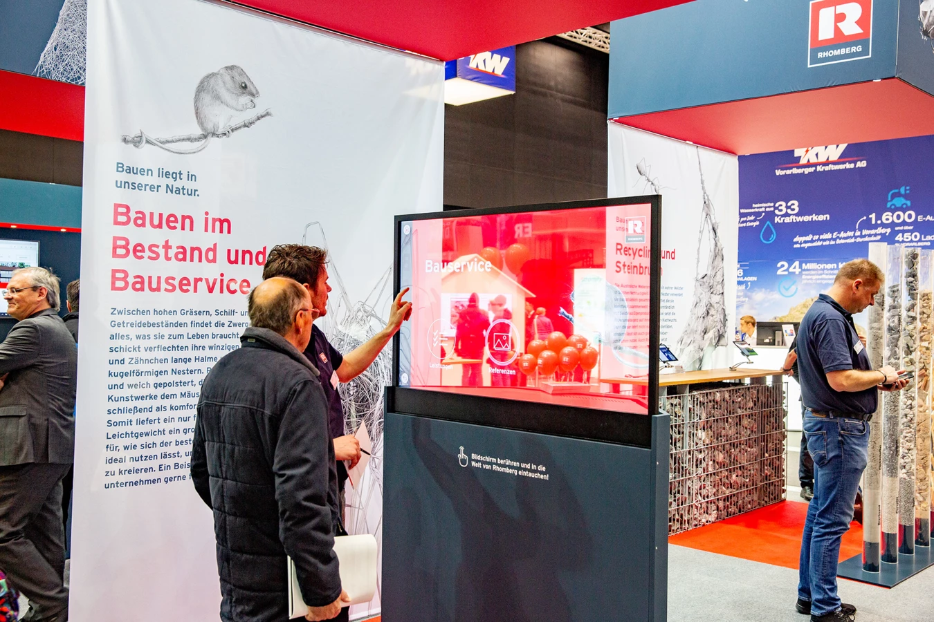 Besucher interagieren mit einem Touchscreen-Display am Rhomberg Bau Messestand, der "Bauservice" und "Recycling und Steinbruch" präsentiert.
