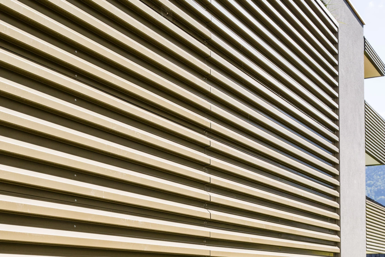 Seitenansicht einer modernen beige-braunen Fassade mit horizontalen Lamellen und vertikalen betongrauen Elementen. Teil der Wohnanlage Bahnhofsstrasse Vandans.
