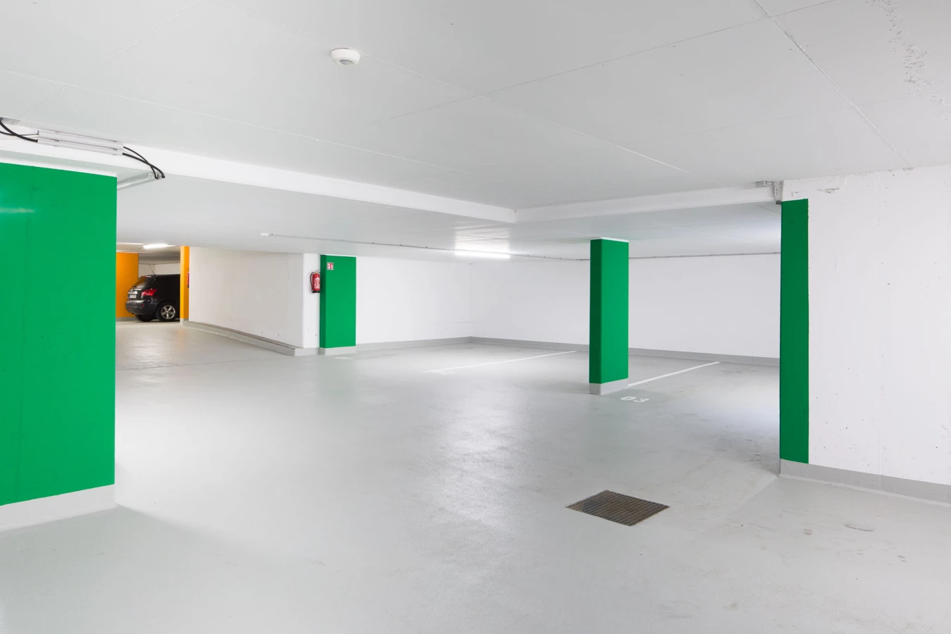 Eine leere, helle Tiefgarage mit grünen Säulen und einem schwarzen Auto am Eingang. Ein Feuerlöscher ist an der Wand sichtbar (Bahnhofstraße, Wohnanlage, Bahnhofsstrasse, Vandans).