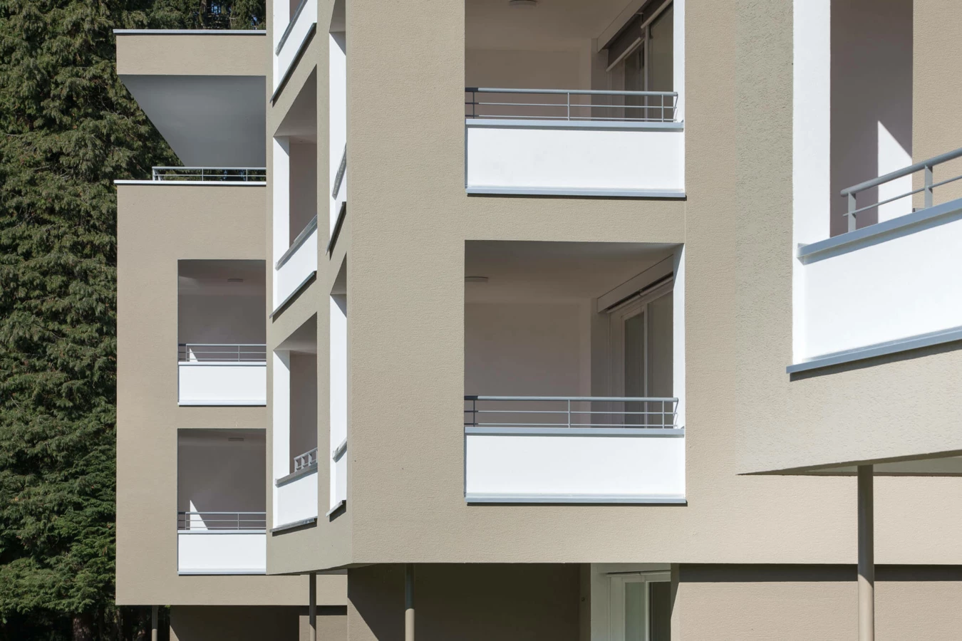 Detailansicht der sandfarbenen Fassade der Wohnanlage Widum Lustenau mit modernen Balkonen und hohen Bäumen im Hintergrund.