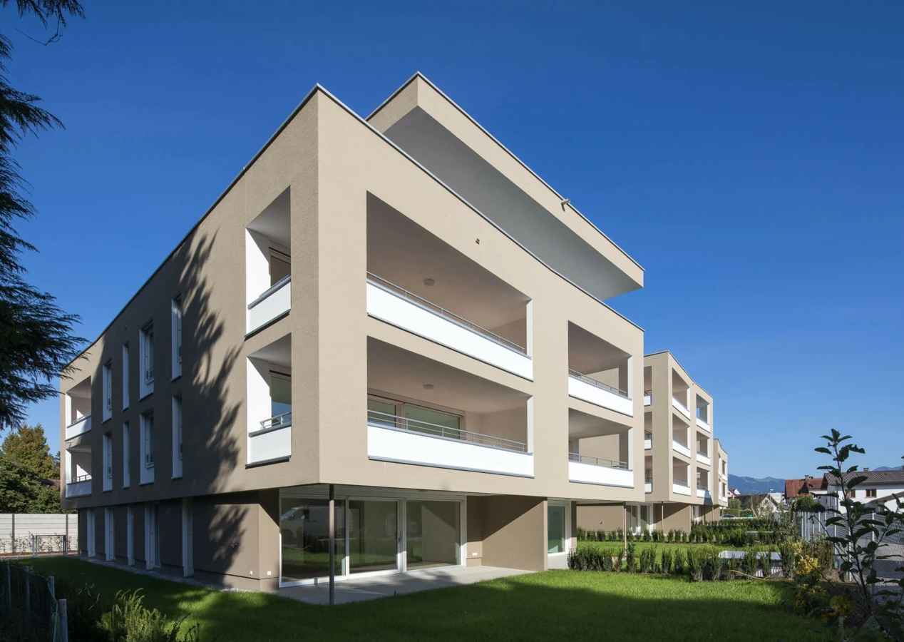 Moderne Wohnanlage Widum in Lustenau mit beigefarbenen Fassaden, mehreren Balkonen und grünen Rasenflächen unter blauem Himmel.