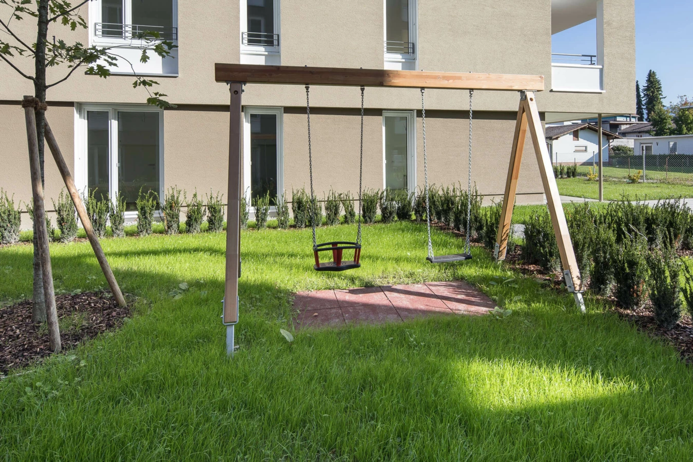 Ein Kinderspielplatz mit Schaukeln auf einer grünen Wiese vor einem modernen Wohngebäude der Wohnanlage Widum Lustenau.