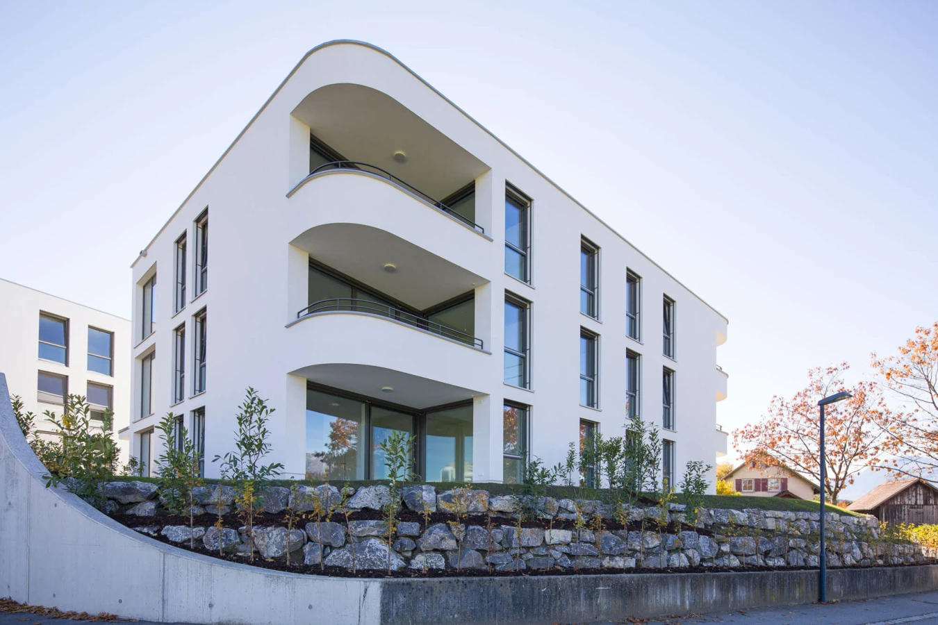 Modernes, weisses Wohngebäude mit geschwungenen Balkonen und grossen Fenstern, eingebettet in eine begrünte Steinmaueranlage (Wolf-Huber-Straße, Wohnanlage, Feldkirch).