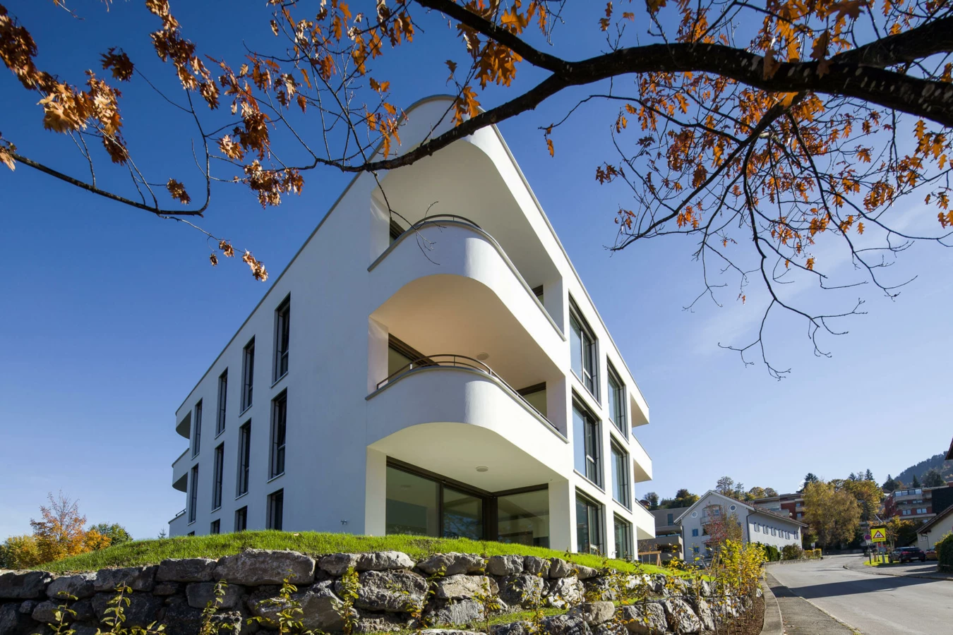 Modernes, weisses Mehrfamilienhaus mit runden Balkonen und grossen Fenstern an der Wolf-Huber-Straße in Feldkirch unter blauem Himmel mit Herbstlaub (Wohnanlage).