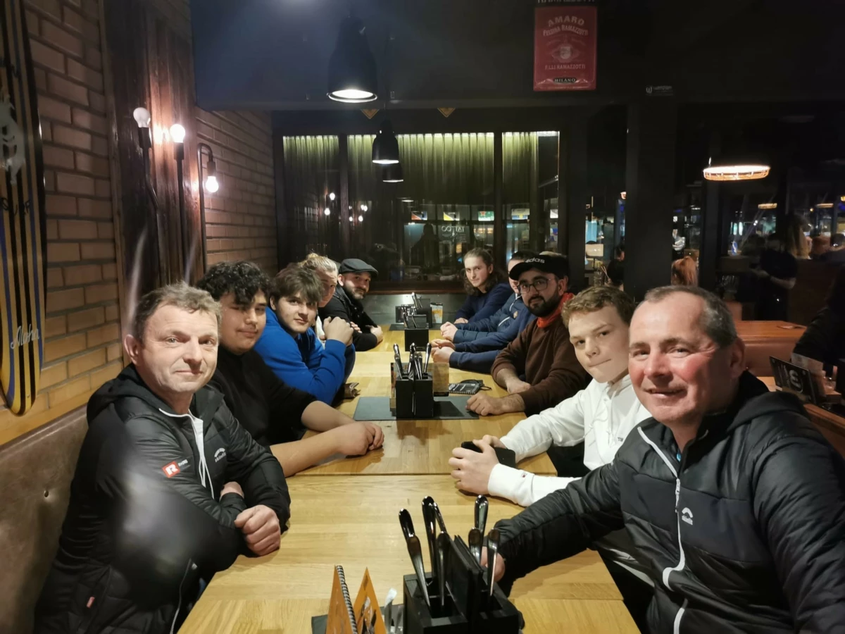 Eine Gruppe junger Männer und zwei ältere Männer sitzen gemeinsam an einem langen Holztisch in einem Restaurant und schauen in die Kamera (Lehrlingsabend).