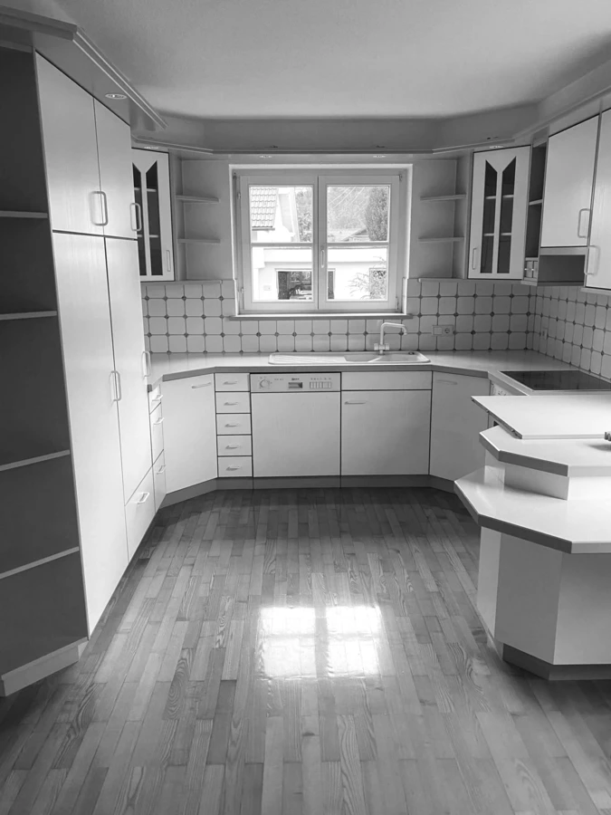 Eine leere, unrenovierte Küche mit weißen Schränken, einer Spüle, einem Geschirrspüler und Holzfußboden, aufgenommen vor der Generalsanierung Rhomberg Bau EFH Bohle.