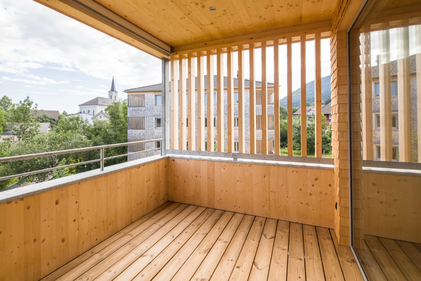 Blick von einem holzverkleideten Balkon mit Holzlatten auf eine Wohnanlage in Alberschwende und einen Kirchturm