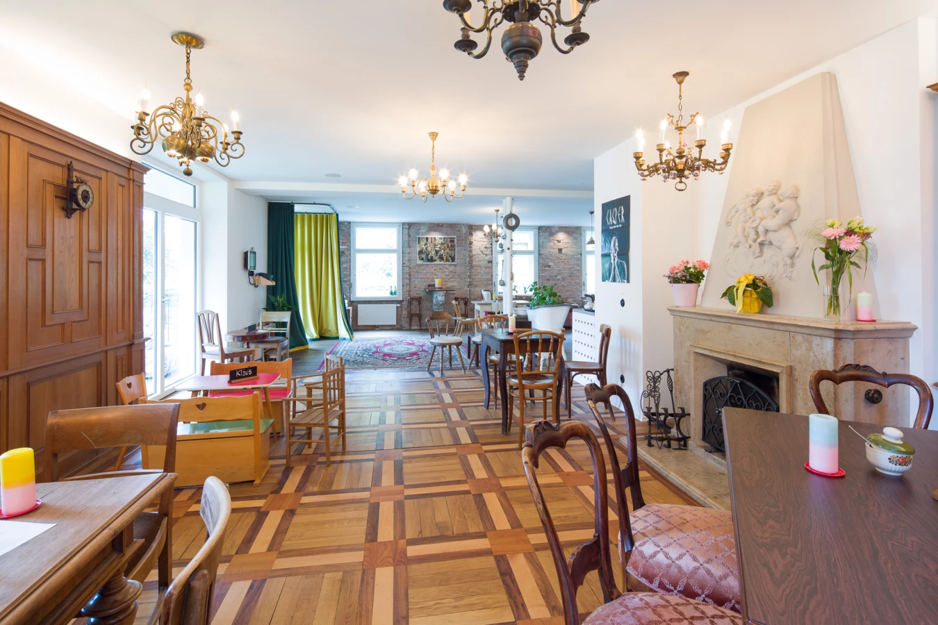 Ein geräumiges Interieur mit einem Fischgrätparkettboden, Kronleuchtern, Tischen und Stühlen, einem hellen Kamin und großen Fenstern mit Vorhängen (Wunderkind, Backstube).