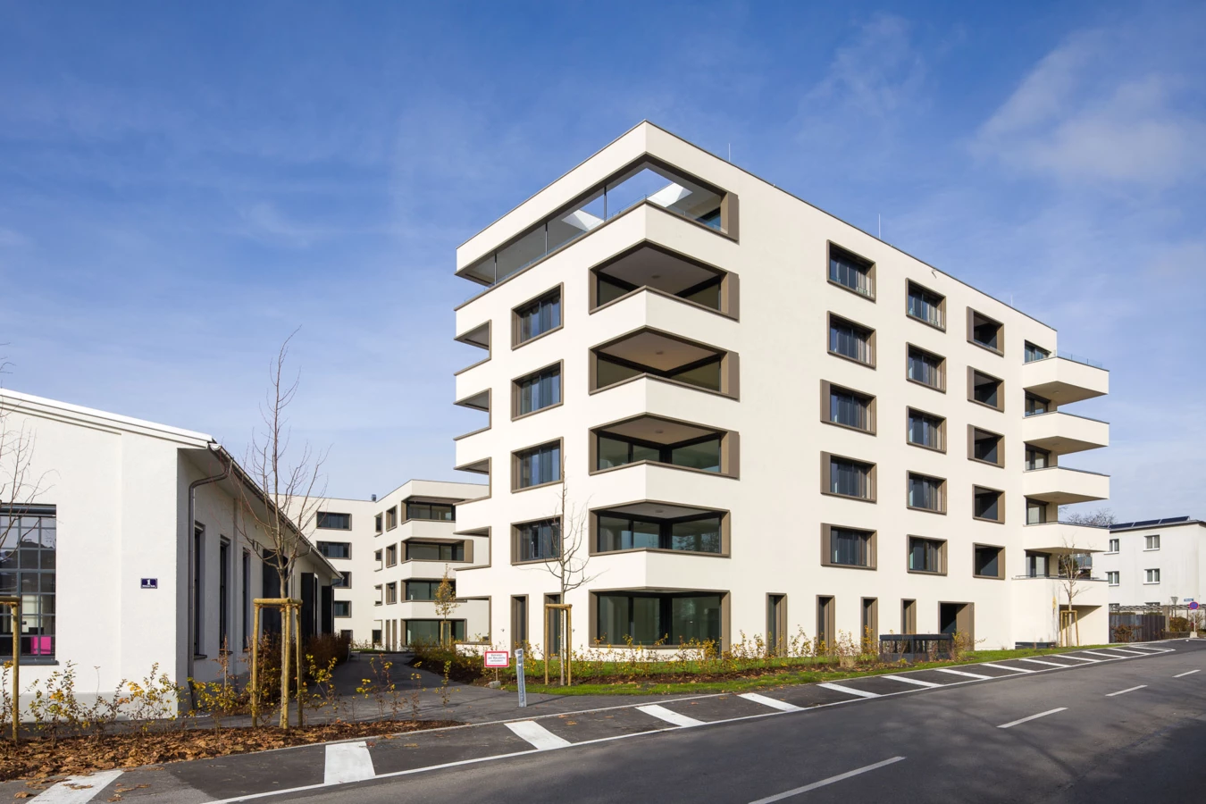 Moderne, weiße Mehrfamilienhäuser mit Balkonen und großen Fenstern unter blauem Himmel, mit einer Straße im Vordergrund (Bäumleareal).