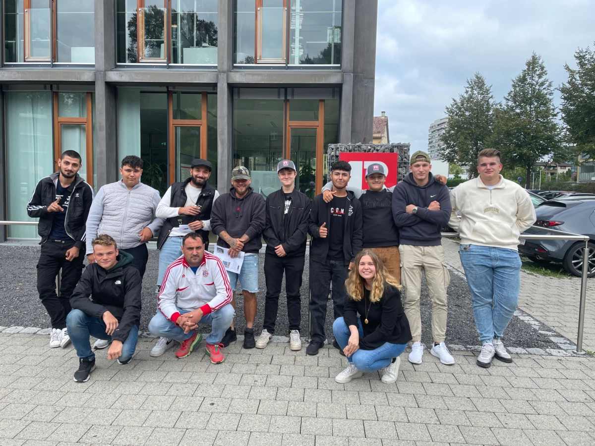 Eine Gruppe junger Männer und eine junge Frau posieren vor einem modernen Bürogebäude, teilweise in die Hocke gegangen (Lehre).