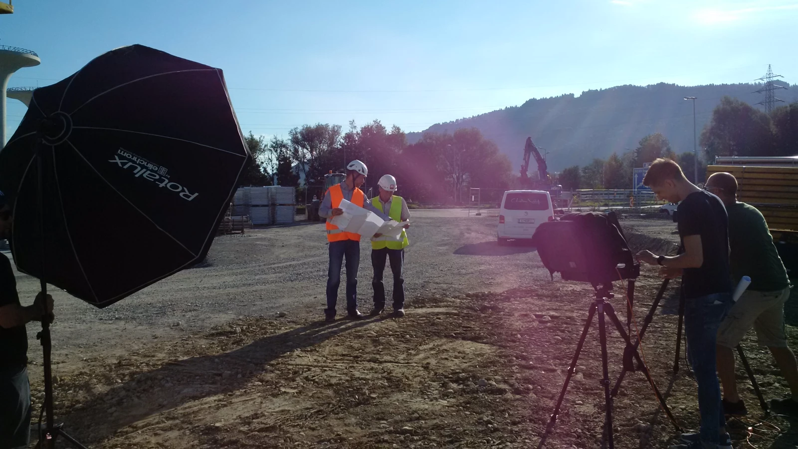 Zwei Bauarbeiter in Warnwesten und Helmen prüfen Baupläne auf einer Baustelle, während ein Fotograf filmt.