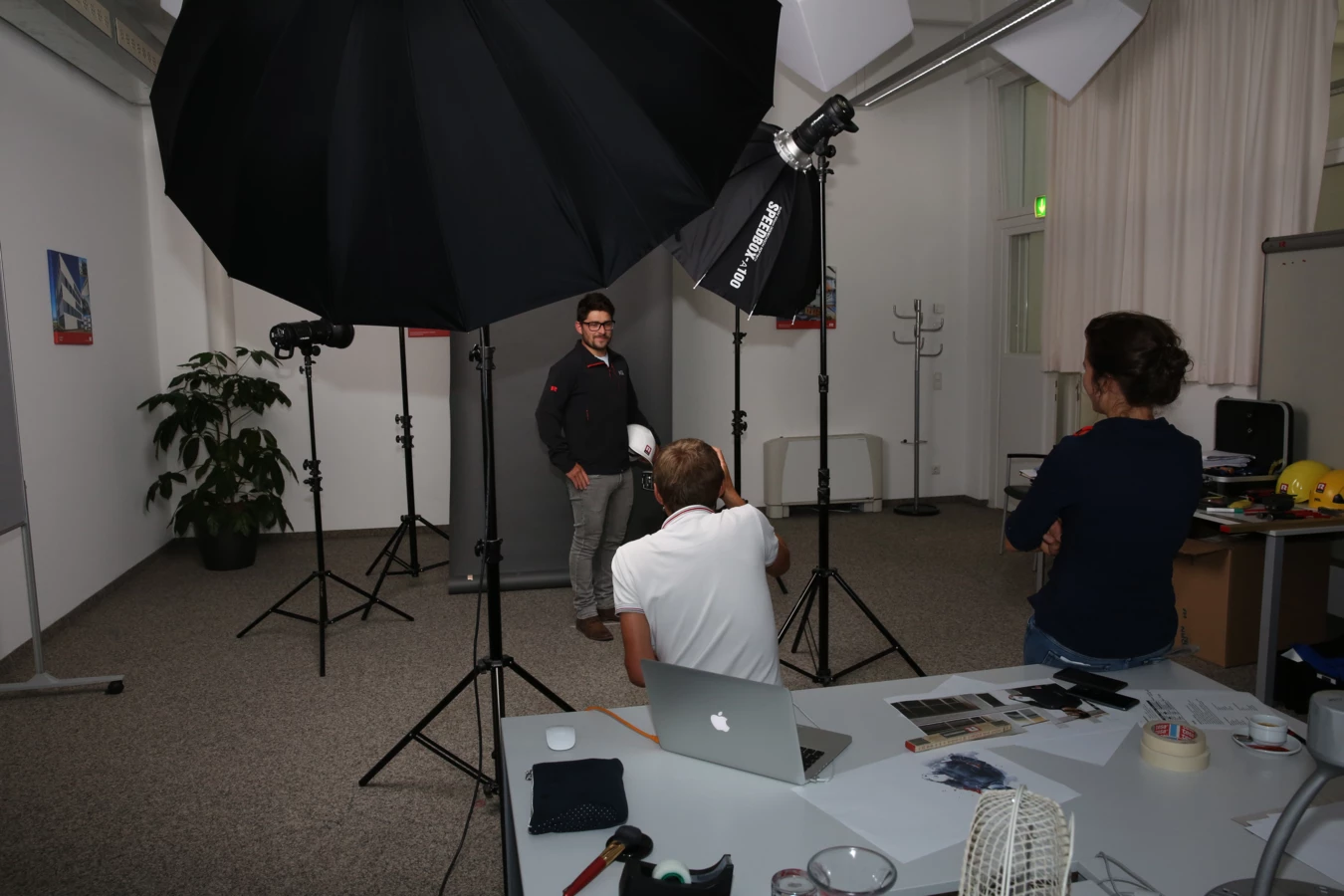 Ein Mann posiert für ein Fotoshooting mit Blitzgeräten und Regenschirmen, während ein Fotograf und ein anderer Mann zusehen.