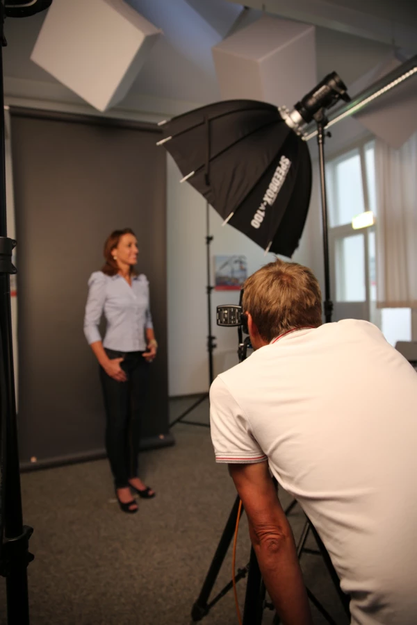 Ein Fotograf macht Aufnahmen einer Frau mit einer Kamera, umgeben von professioneller Beleuchtung und einem grauen Hintergrund.