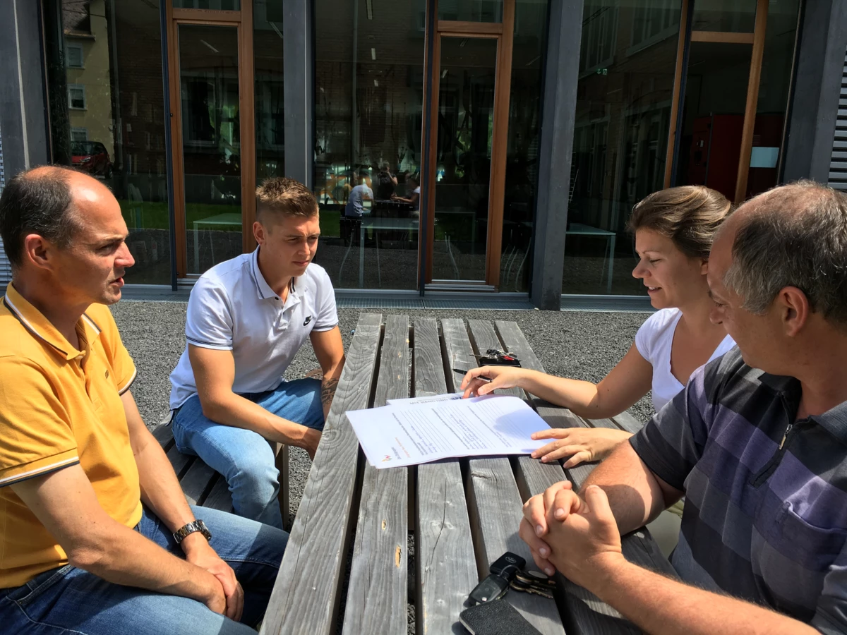 Vier Personen, zwei Männer, eine Frau und ein junger Mann, sitzen an einem Holztisch im Freien und besprechen Dokumente (Workshop).