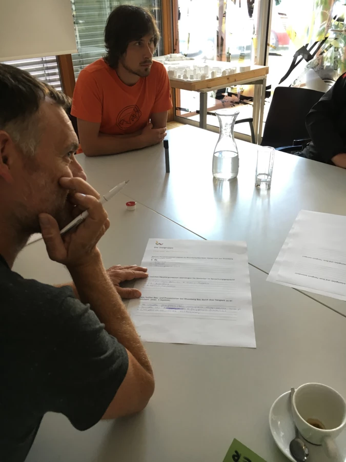Zwei Männer sitzen an einem Tisch, auf dem Papiere, Stifte und eine Kaffeetasse liegen. Im Hintergrund ist ein Architekturmodell sichtbar (Workshop).
