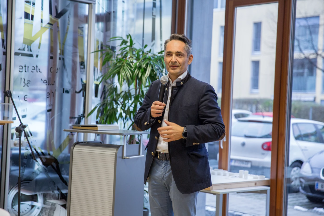 Ein Mann im Sakko spricht vor einem Mikrofon in einem modernen Raum mit Glasfenstern und Ausblick ins Freie (Hubert, Geburtstag).