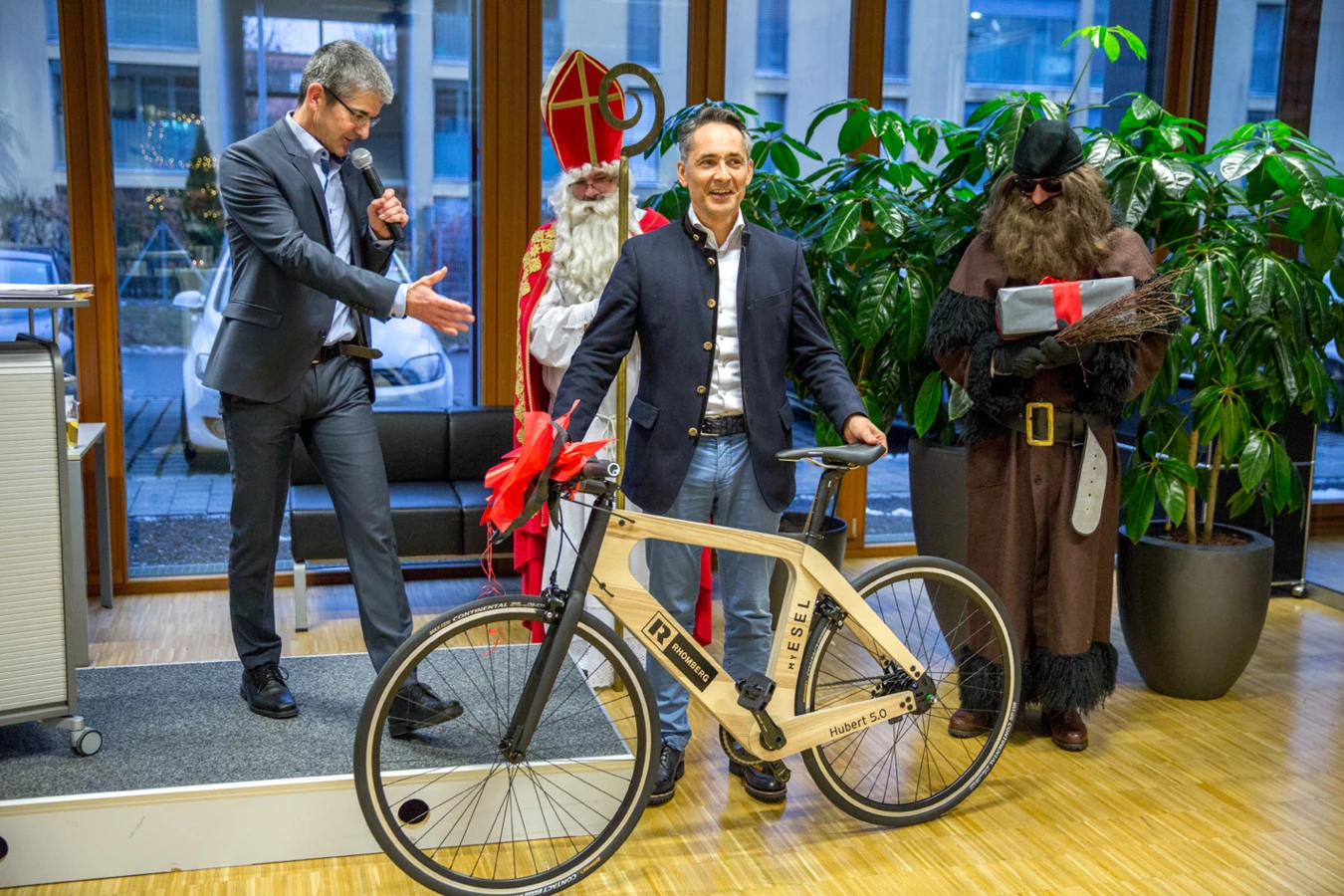 Geschäftsleute und verkleidete Personen präsentieren ein Holzfahrrad Hubert 5.0 mit dem Rhomberg Logo auf einer Bühne (Geburtstag).