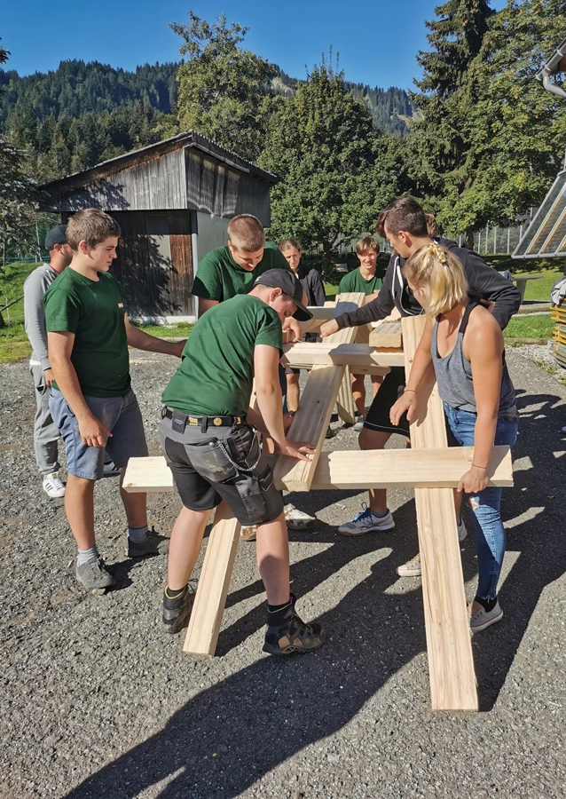 Mehrere Lehrlinge von Rhomberg Bau montieren gemeinsam an einem sonnigen Tag Holzteile im Freien (Zusammenbauen).