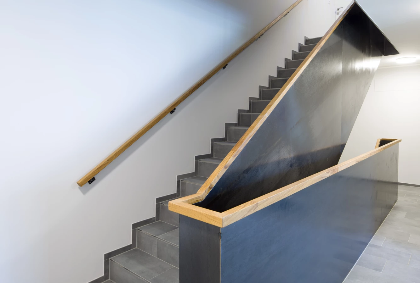 Eine moderne Treppe mit Holzgeländern und dunkelgrauen Metallbrüstungen in einem Gebäude der Unterhochstegstrasse