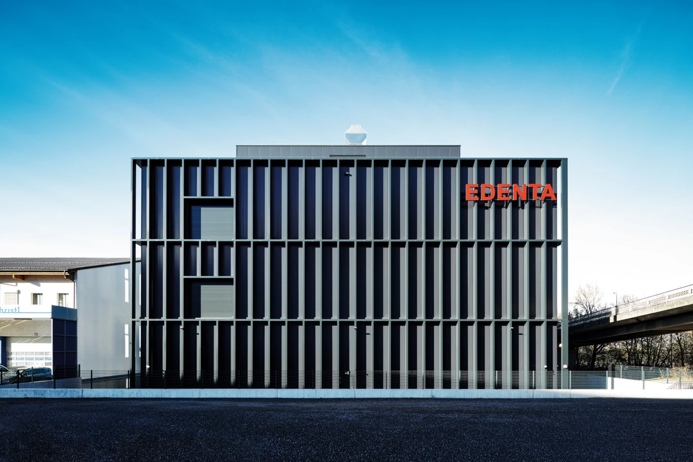 Modernes, quaderförmiges Geschäftsgebäude mit vertikalen Lamellen und Leuchtschrift „EDENTA“ an der Fassade unter blauem Himmel.