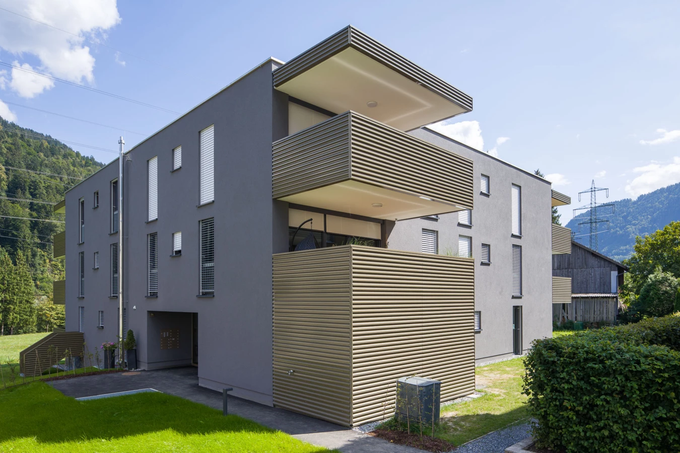 Moderne Wohnanlage in Vandans mit grauer Fassade, Balkonen mit Holzlamellen und grünem Rasen im Vordergrund unter blauem Himmel (Bahnhofstraße, Bahnhofsstrasse)