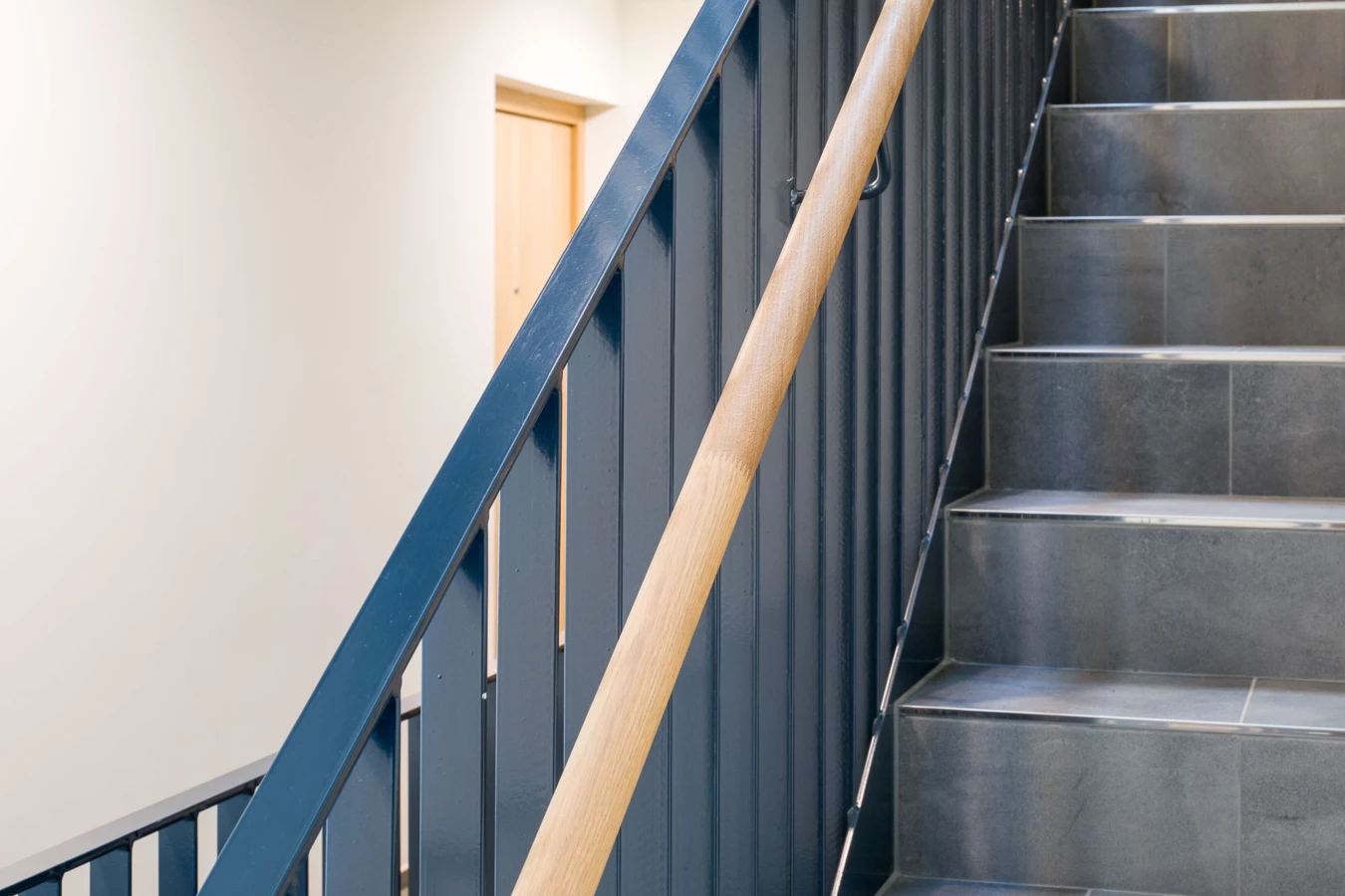 Eine dunkelblaue Metalltreppe mit einem Holzhandlauf und grau gefliesten Stufen in der Wohnanlage Bahnhofsstrasse Vandans.