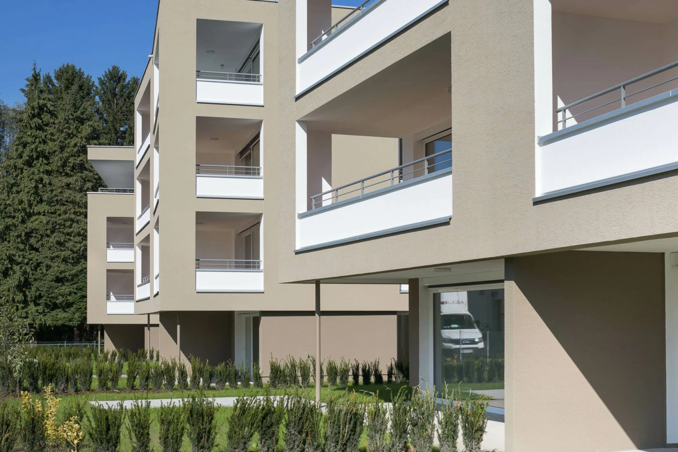 Moderne Wohnanlage Widum Lustenau mit hellgrauer Fassade, weissen Balkonbrüstungen und gepflegter Grünfläche mit Büschen