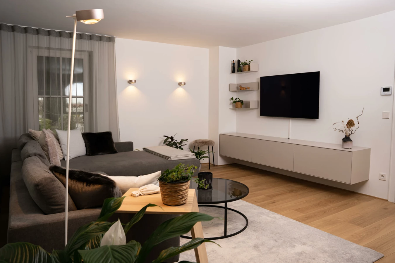 Modernes Wohnzimmer mit grauem Ecksofa, hellem Holzboden, TV, Wandleuchten und Zimmerpflanzen (Generalsanierung, Bohle)