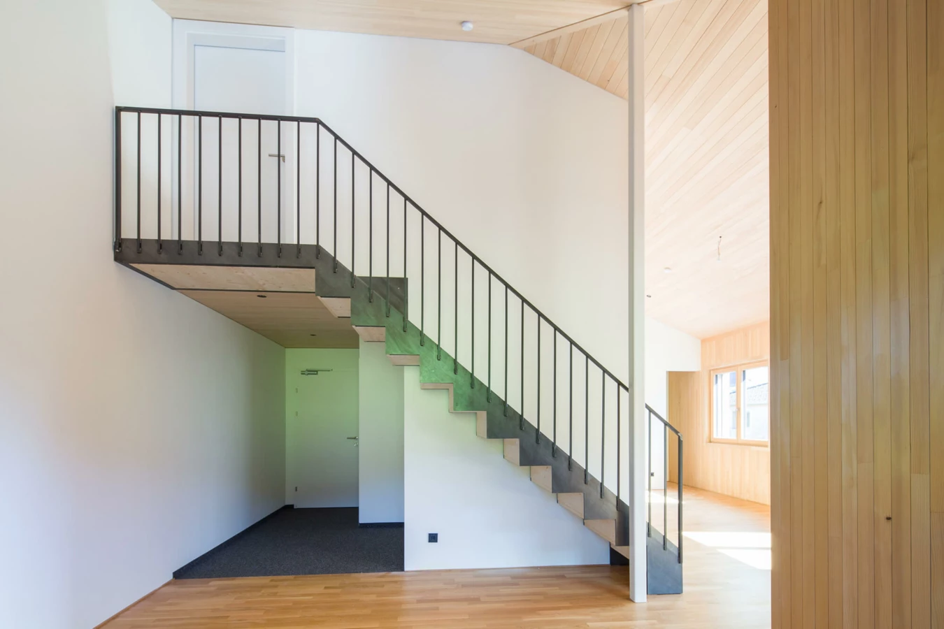 Eine moderne Holztreppe mit schwarzem Metallgeländer in einem hellen Eingangsbereich mit Holzboden und weißen Wänden (Wohnanlage, Alberschwende).