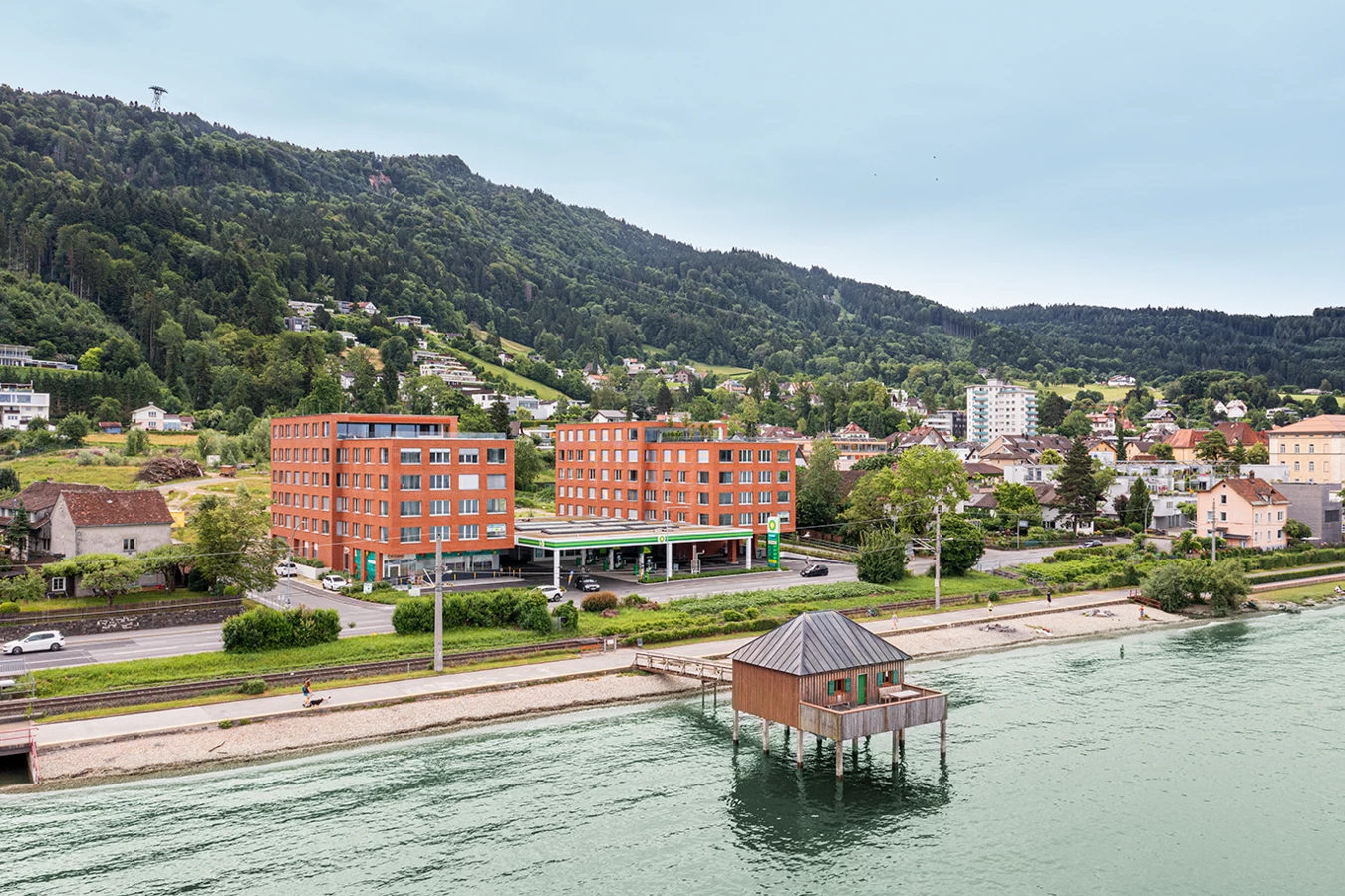 Zwei moderne rote Backsteingebäude neben einer BP-Tankstelle an einer Straße am Bodensee. Ein Holzhaus auf Stelzen ragt ins Wasser, dahinter ein bewaldeter Berg (Seeblick, Lochau).