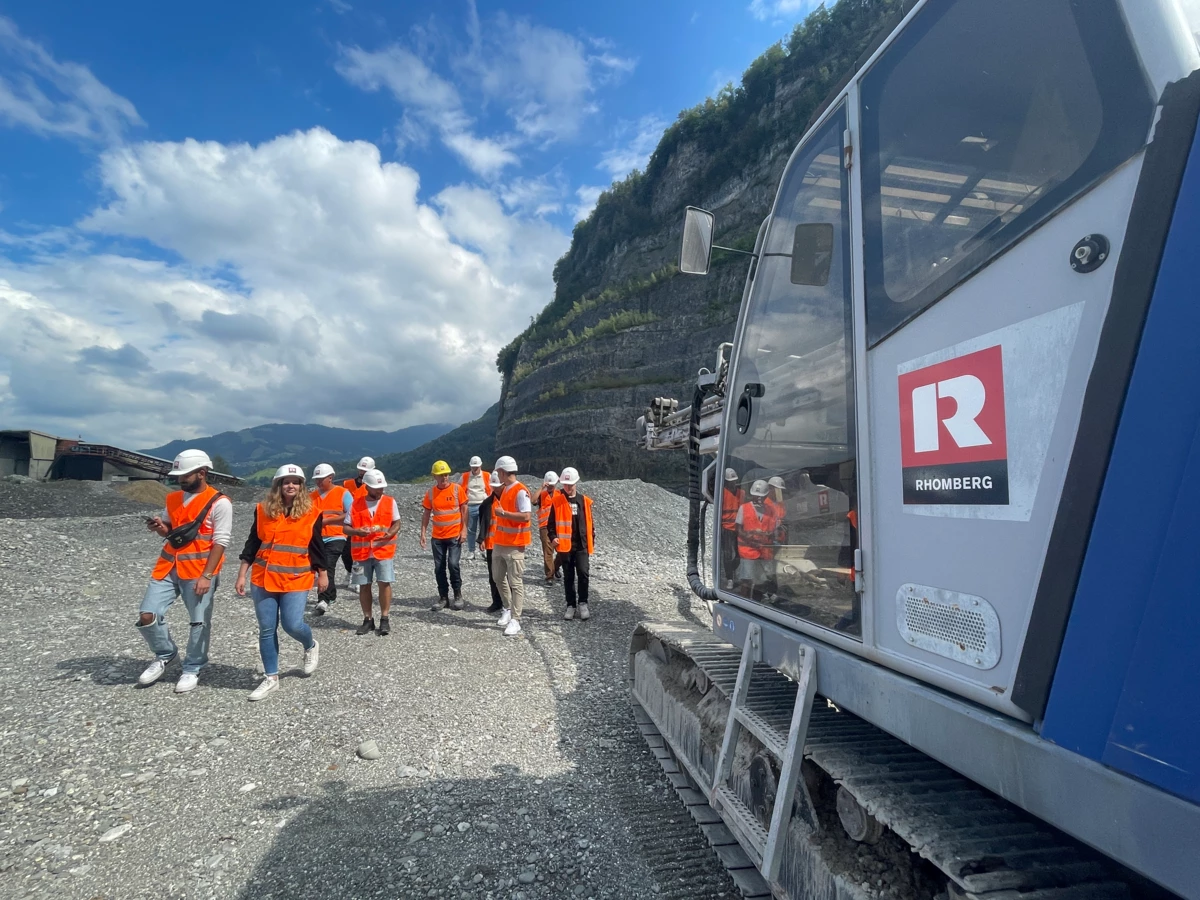 Eine Gruppe junger Menschen in orangefarbenen Westen und Schutzhelmen geht an einem Bagger mit Rhomberg-Logo vorbei auf einem Kiesweg, mit Bergen im Hintergrund (Lehre).