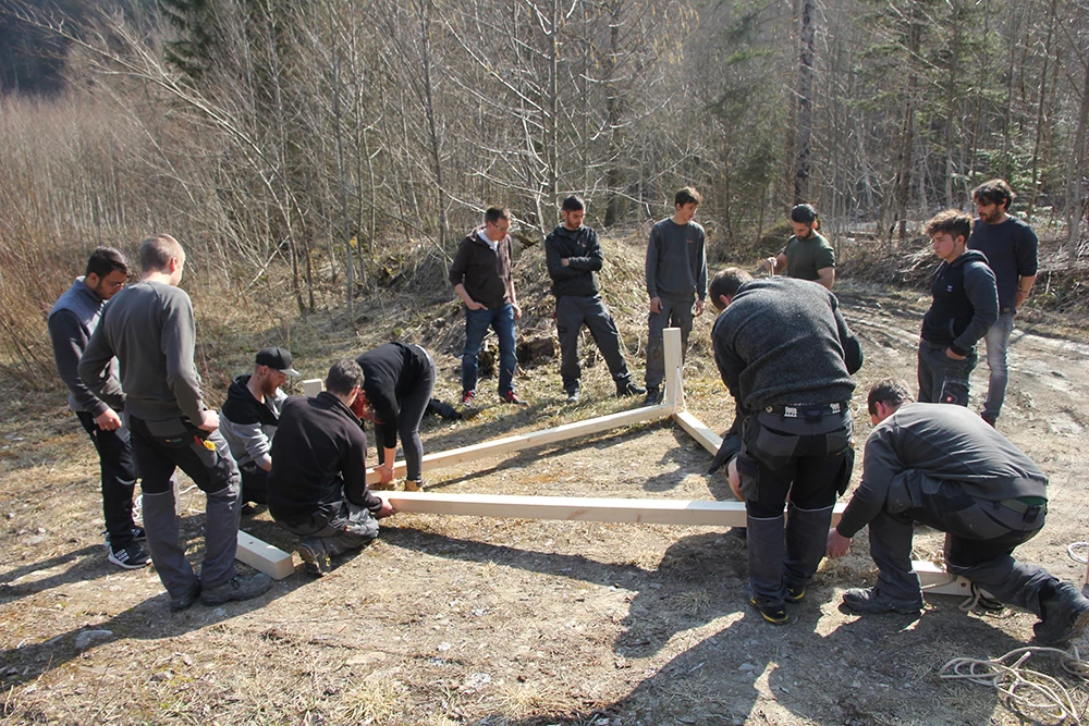 Eine Gruppe junger Männer arbeitet im Freien an einer Holzkonstruktion, umgeben von Bäumen und Sträuchern (Lehrlingstag).