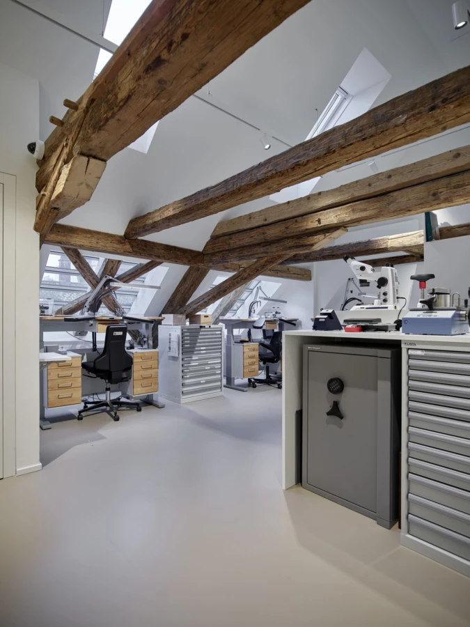 Uhrmacherwerkstatt unter dem Dachgiebel mit Holzbalken. Mehrere Arbeitsplätze mit Mikroskopen, Werkzeugen und Lagerregalen (Huber, Rathausstr).