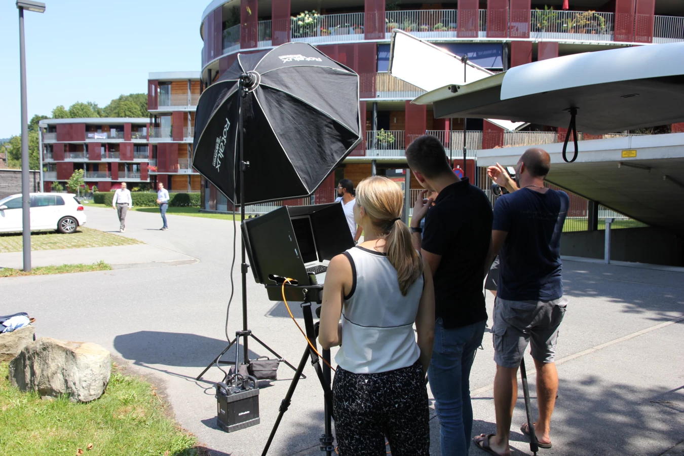 Team bei einem Fotoshooting im Freien, mit Kameraausrüstung, Stativen und Softboxen vor einem modernen Gebäude.
