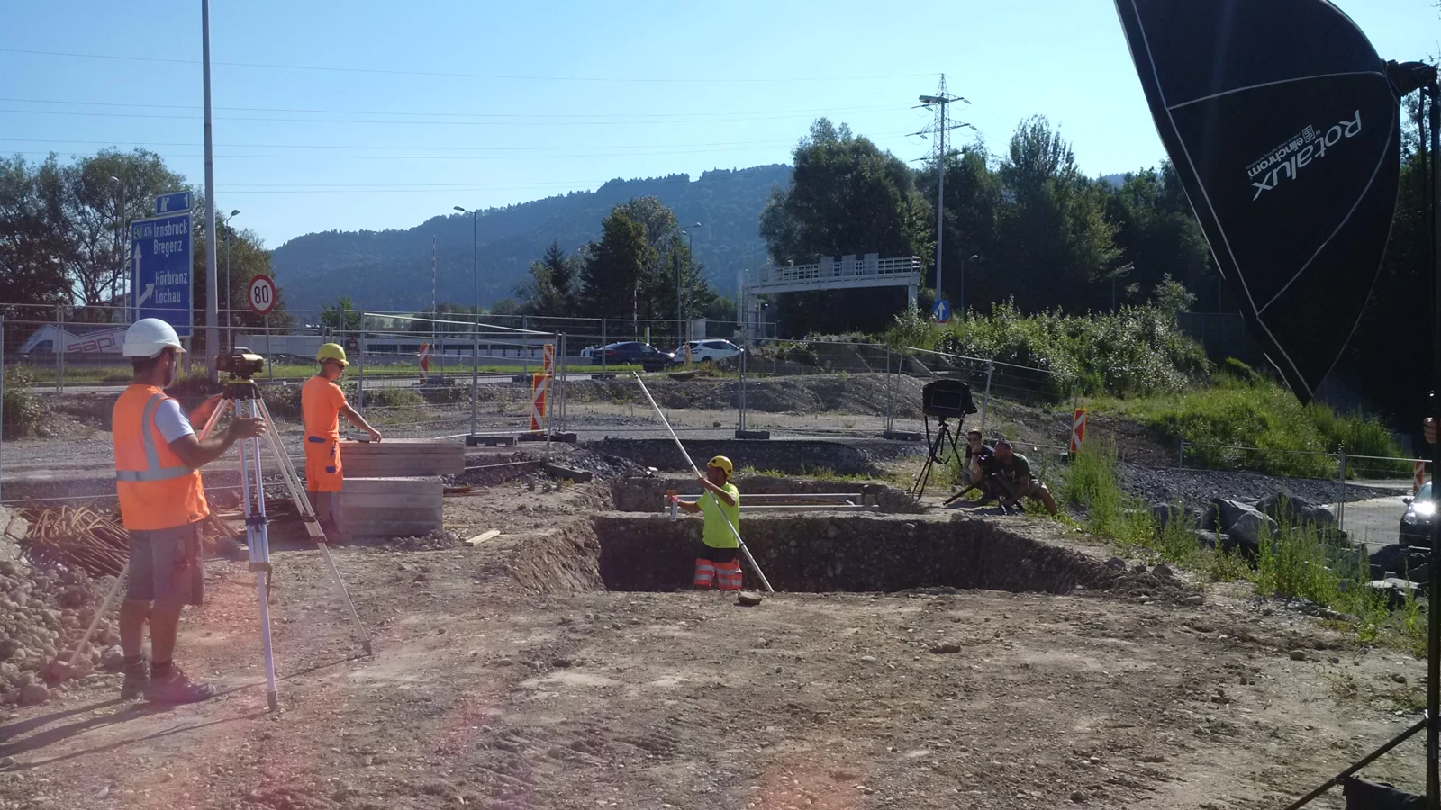 Rhomberg Bau Mitarbeiter auf einer Baustelle mit Vermessungsgeräten und Kamera-Equipment unter blauem Himmel.