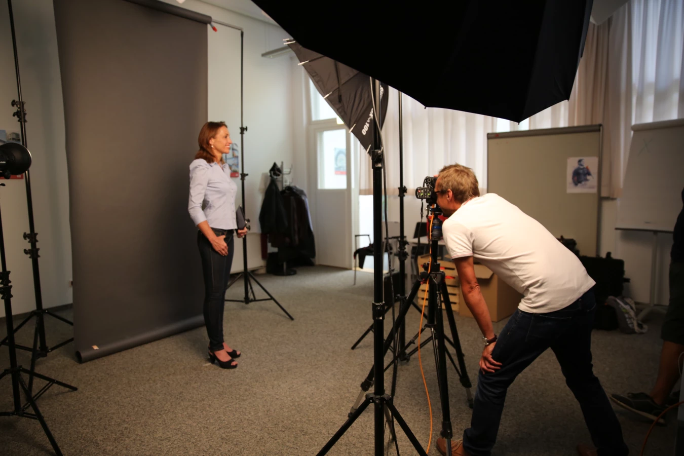 Ein Fotograf macht ein Porträtfoto einer Frau in einem Fotoshooting-Studio mit grauer Kulisse und Beleuchtungsausrüstung.
