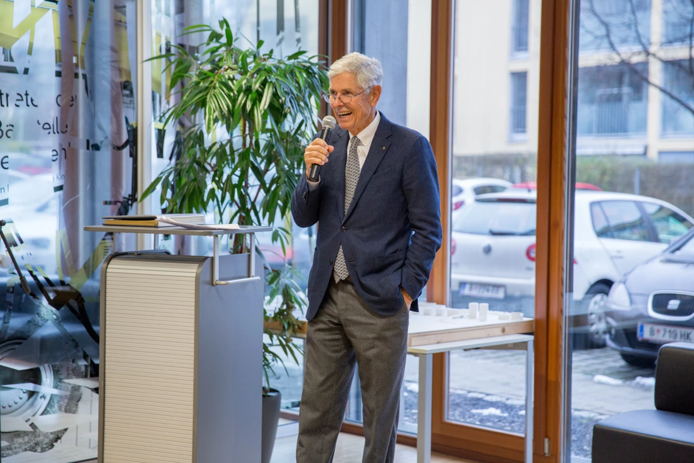 Ein lächelnder, grauhaariger Mann im Anzug hält ein Mikrofon und spricht auf einer Veranstaltung vor großen Fenstern (Hubert, Geburtstag).