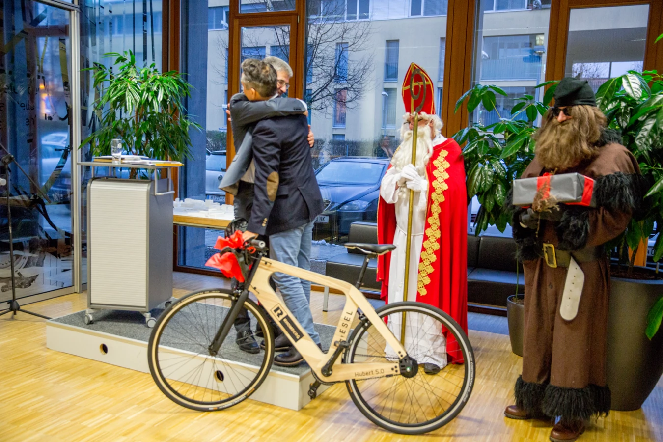 Zwei Männer umarmen sich neben einem hölzernen Rhomberg Fahrrad, während Nikolaus und Krampus bereitstehen (Hubert, Geburtstag).